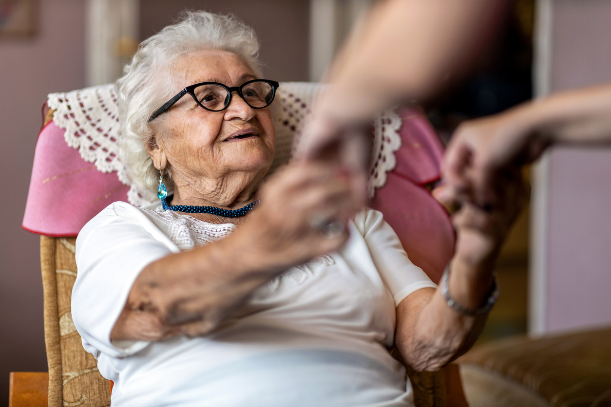 female home carer supporting old woman to stand up from the armchair at care home